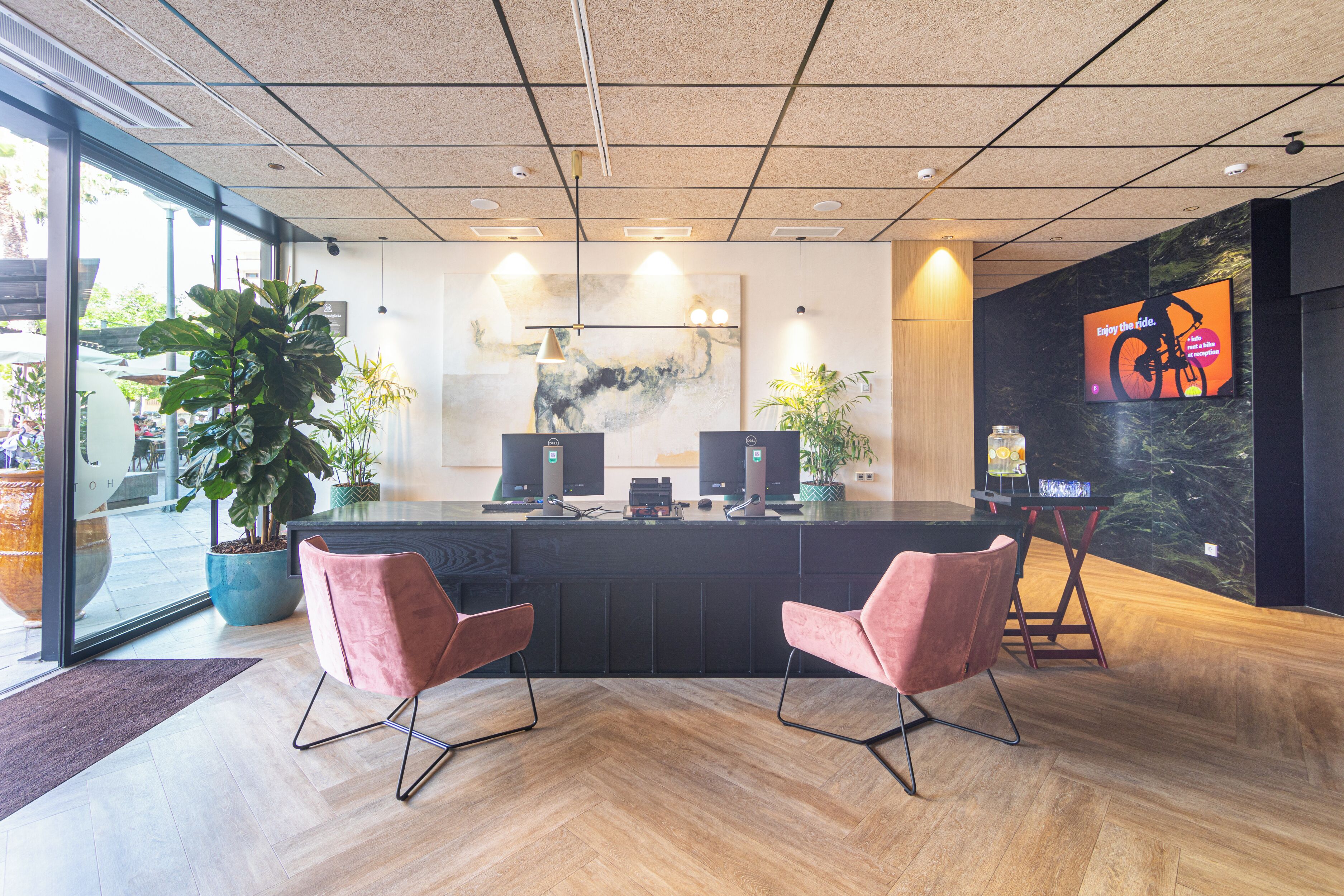 A reception area with a modern design, featuring a reception desk, two pink chairs, a large potted plant, and a mounted television displaying a cycling advertisement.
