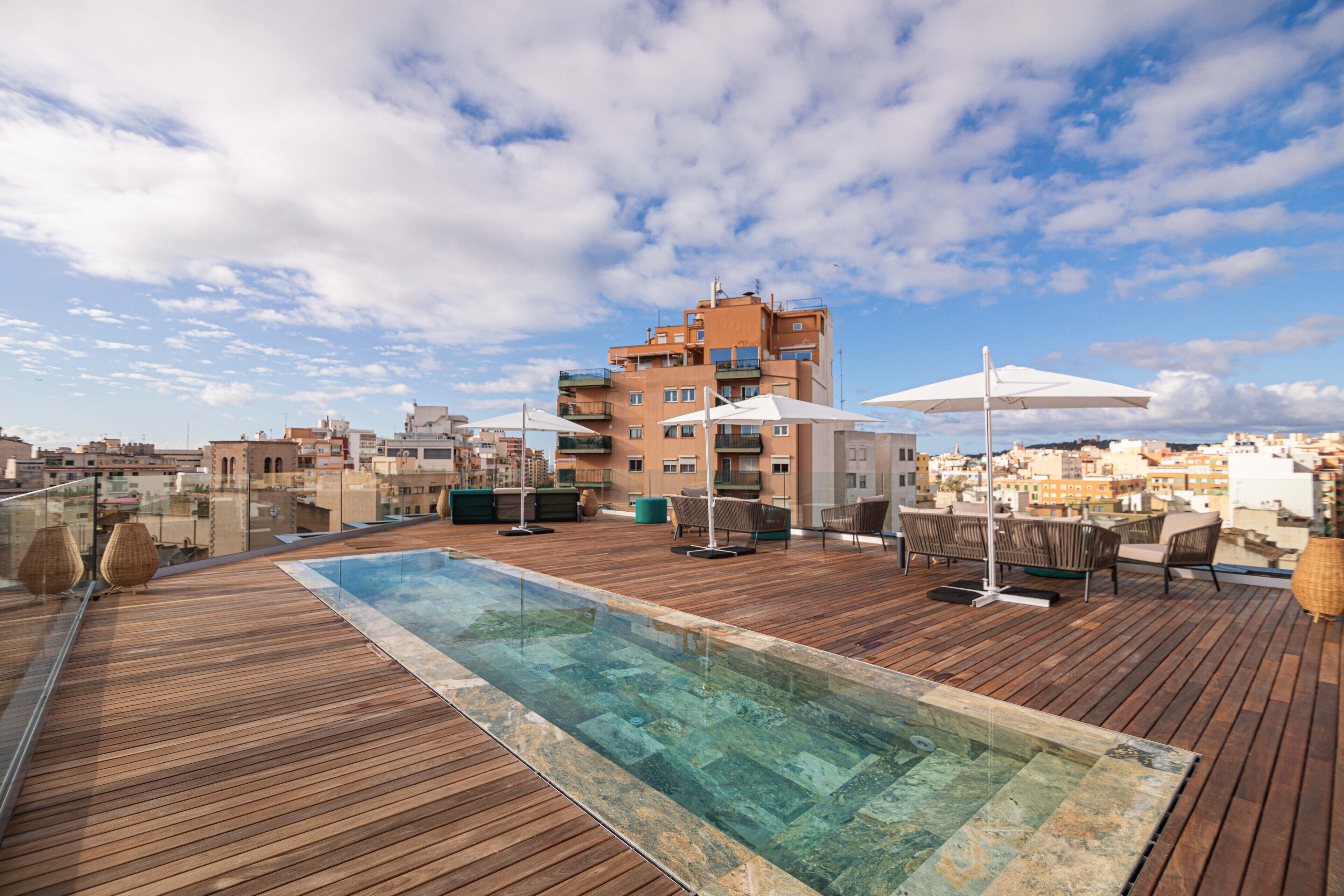 A rooftop pool area with wooden decking, lounge chairs, and umbrellas, overlooking a cityscape with buildings and a clear sky.