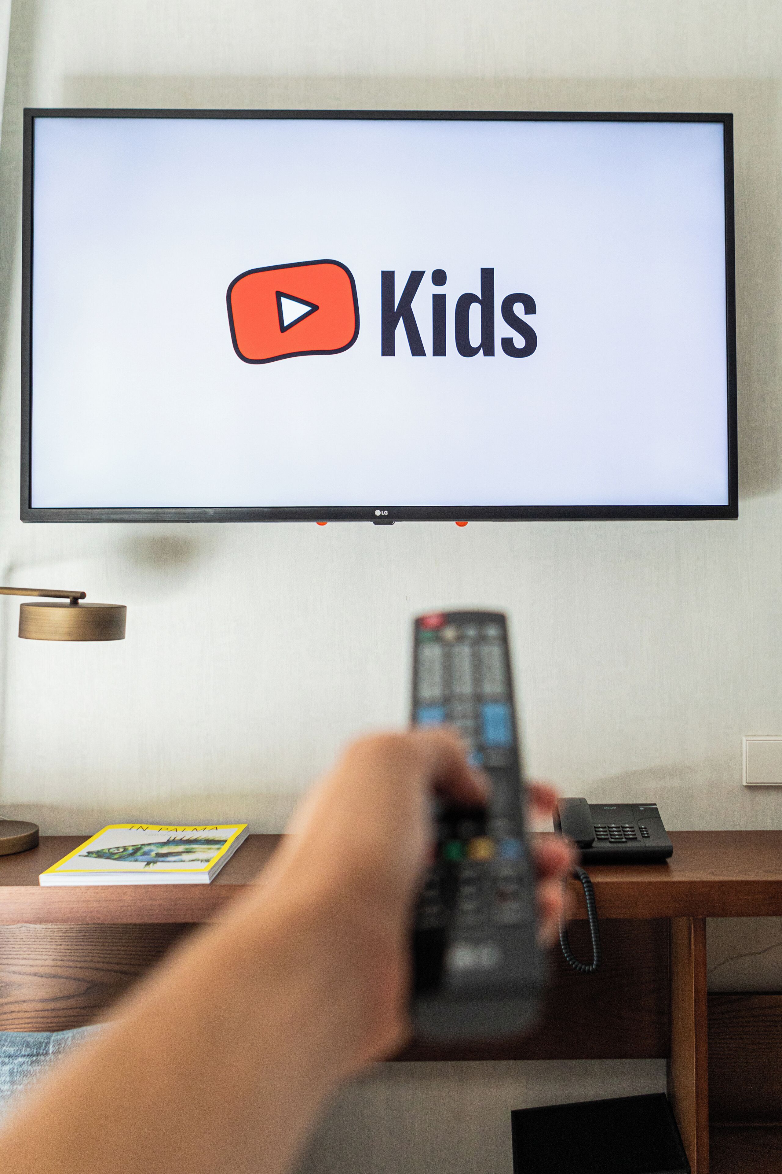 A television displaying the YouTube Kids logo, a remote control in hand, and a book titled "IN BETA" on a wooden table.