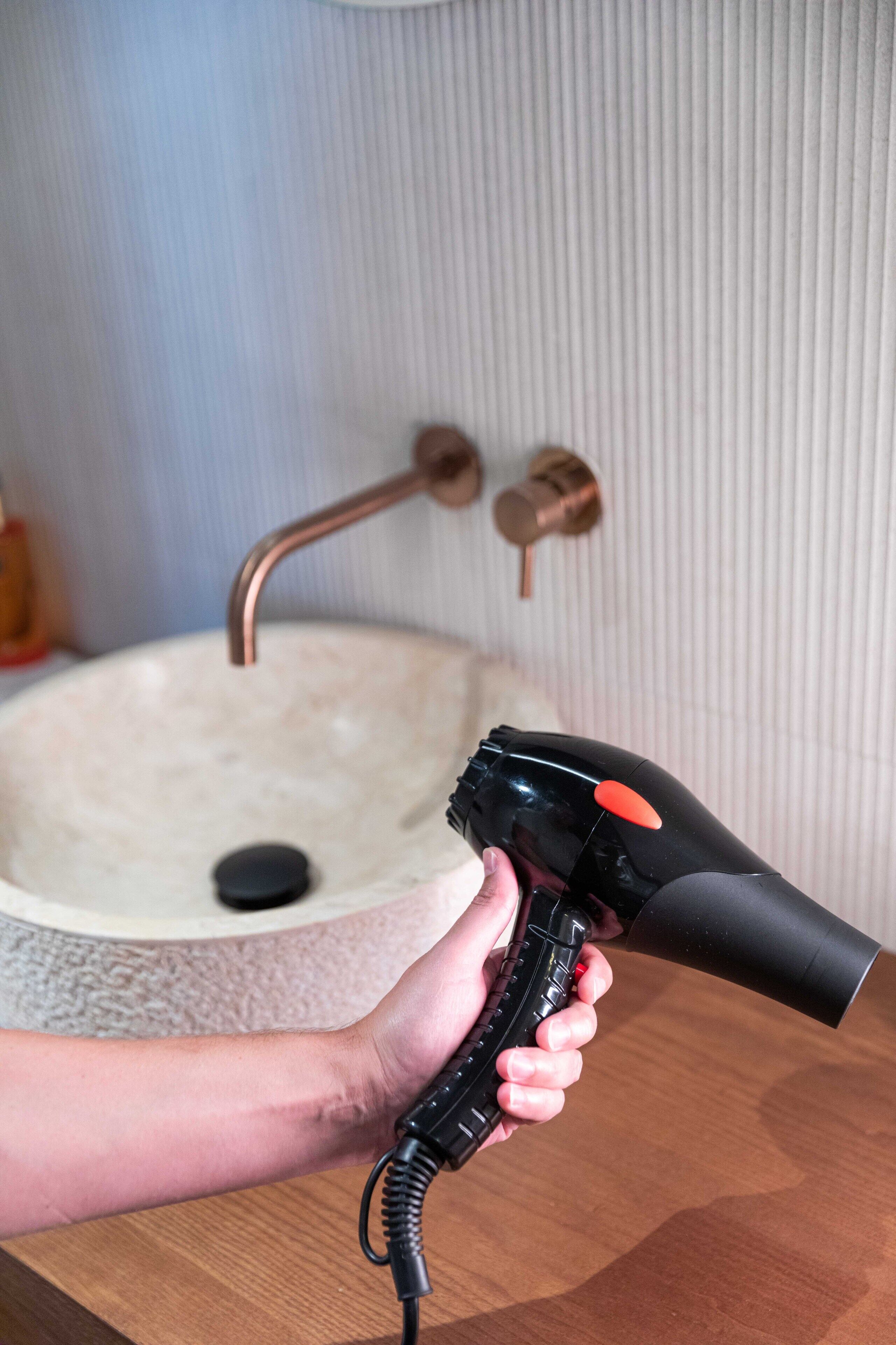 Une main tenant un sèche-cheveux près d’un lavabo dans une salle de bain, avec un robinet en cuivre.