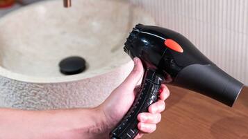 Une main tenant un sèche-cheveux près d’un lavabo dans une salle de bain, avec un robinet en cuivre.