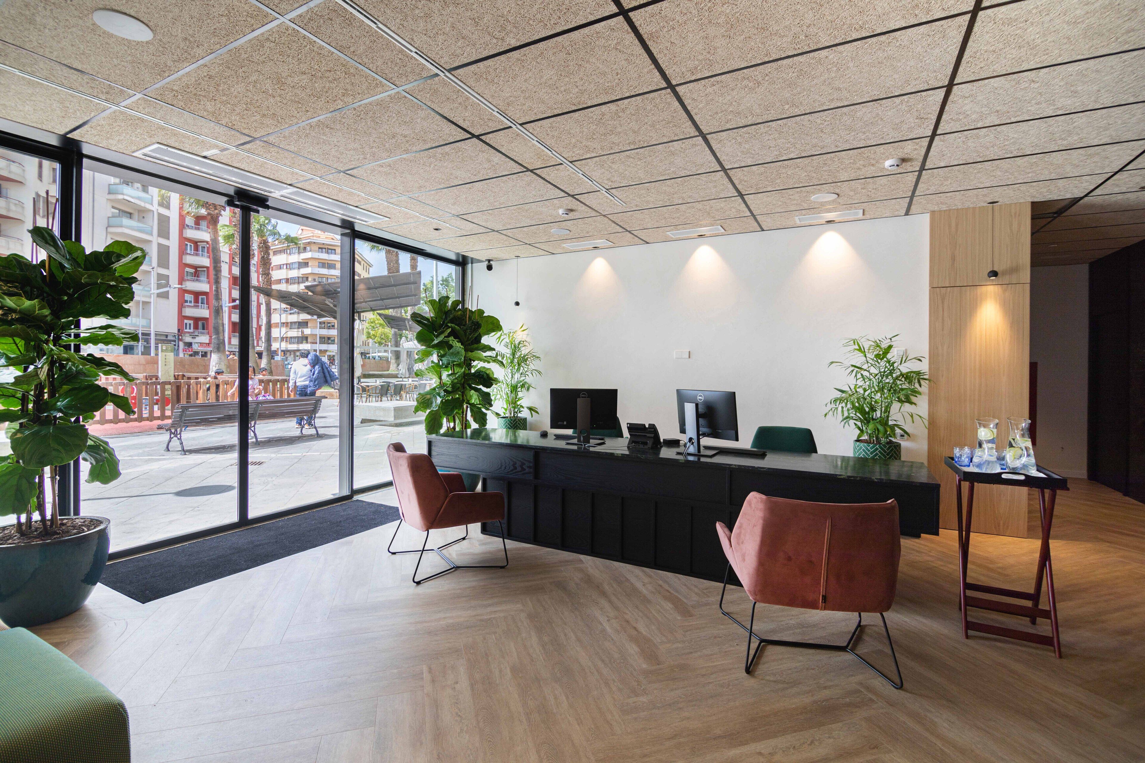 Un espacio de oficina moderno con un mostrador de recepción, dos sillas, plantas en macetas y vistas de la ciudad a través de grandes ventanales.