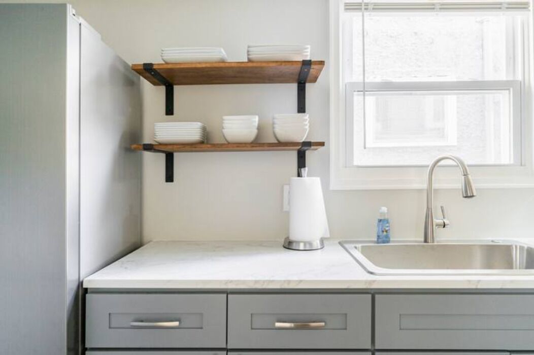 A kitchen with a white countertop, a stainless steel sink, and wooden shelves above it.