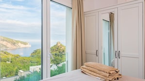 A bedroom with a large window offering a scenic view of a coastline and greenery.