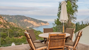 An outdoor wooden deck with a round table and four chairs, overlooking a coastal landscape with hills and the sea.