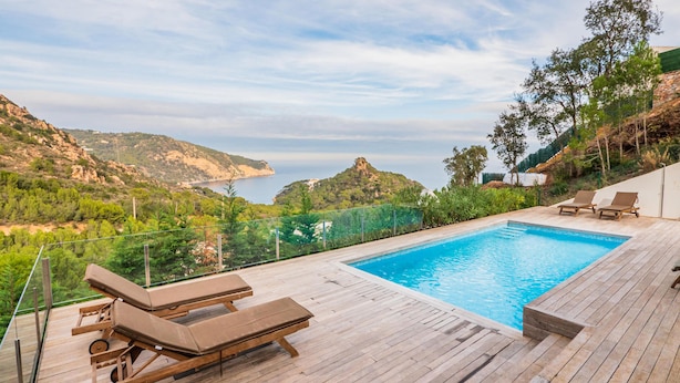 A wooden deck with a swimming pool, lounge chairs, and a scenic view of the sea and hills.