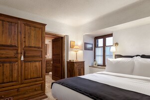 Un dormitorio con un amplio armario de madera, una cama con ropa blanca, una mesita de noche con lámpara y una ventana con cortinas.