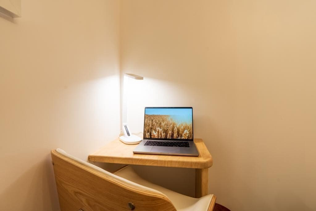 Una laptop sobre un escritorio de madera con una lámpara blanca, una silla de madera y una pared lisa.