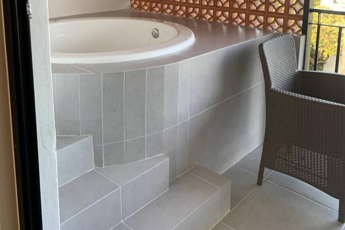 Un baño con bañera, escalones de cerámica, una silla de mimbre y un panel de madera decorativo.