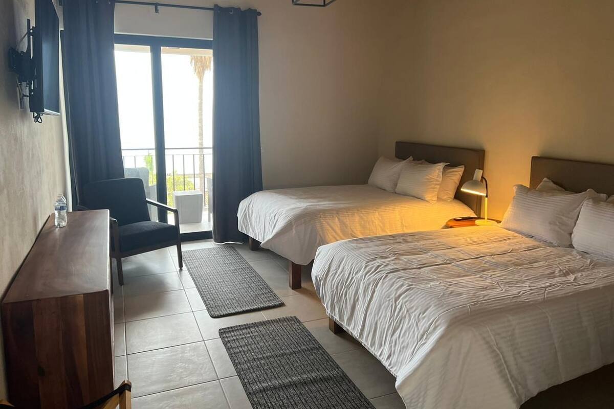 Una habitación de hotel con dos camas, un escritorio, una silla y un balcón con cortinas.