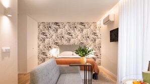 Una habitación de hotel moderna con sofá, una mesita, cama con cabecera, televisor montado en la pared y ventana con cortinas.