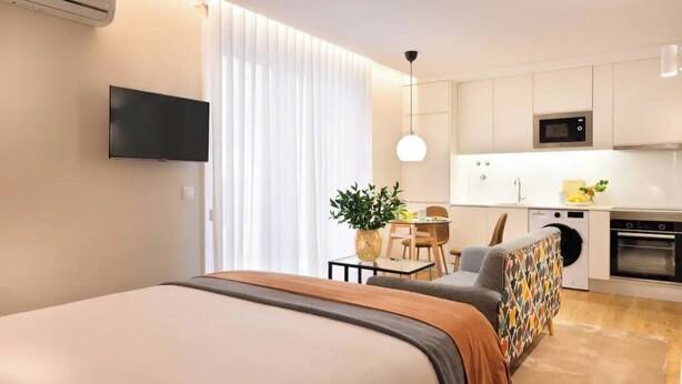 Una habitación de hotel moderna con cama, televisor, comedor con mesa y sillas, cocina con microondas y horno, y un amplio ventanal con cortinas.