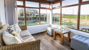 A sunlit room with large windows, a sofa, a table, and a view of a green landscape.