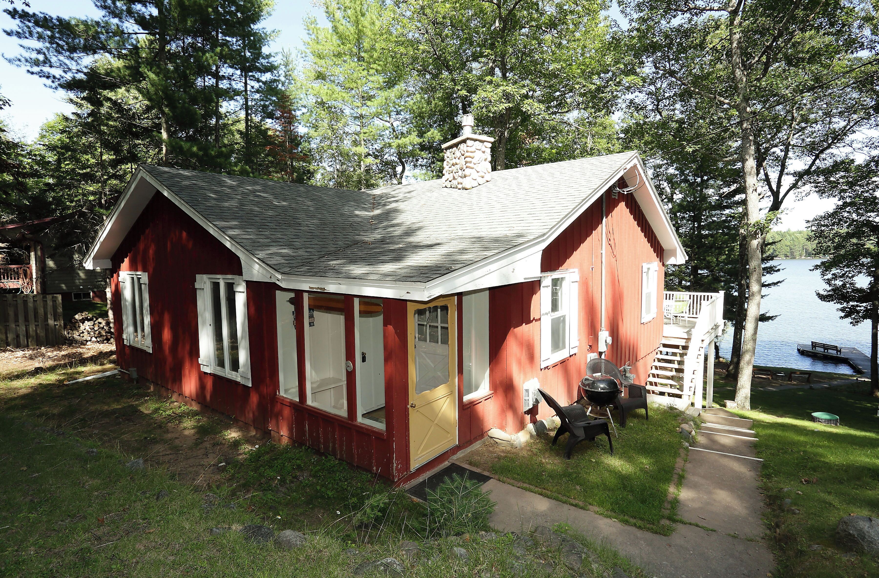 A red cabin with a chimney, large windows, and a dock by a lake.