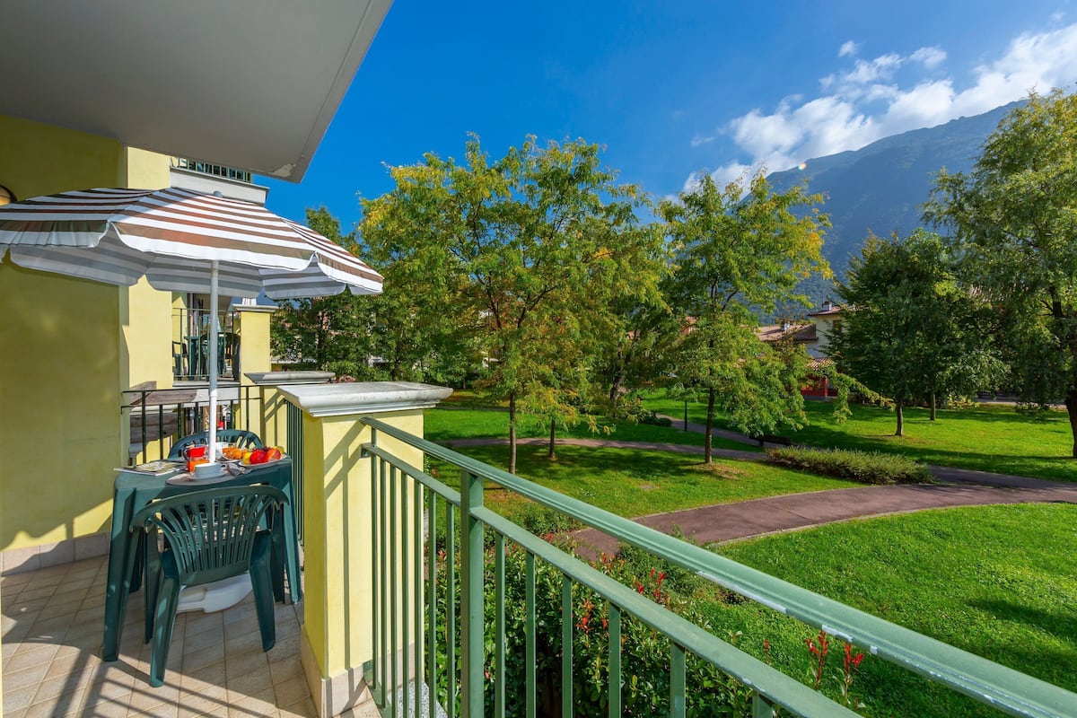 Ein Balkon mit Tisch und Stühlen, einem gestreiften Sonnenschirm und Blick auf eine grüne Rasenfläche und Bäume.