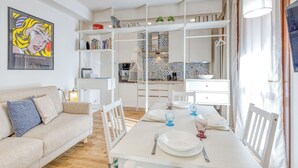 Una sala de estar moderna y bien iluminada con comedor, muebles blancos y una cocina al fondo.