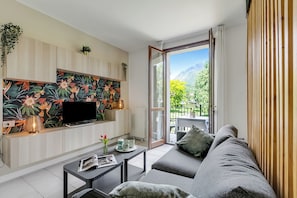 A modern living room with a flat-screen TV, a sofa, a coffee table, and a balcony with a mountain view. - Village Apartments - Porto Letizia (Porlezza)