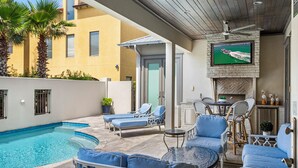 Un espace aménagé au bord de la piscine, avec des chaises longues, un téléviseur fixé sur un mur en pierre et un ventilateur de plafond.