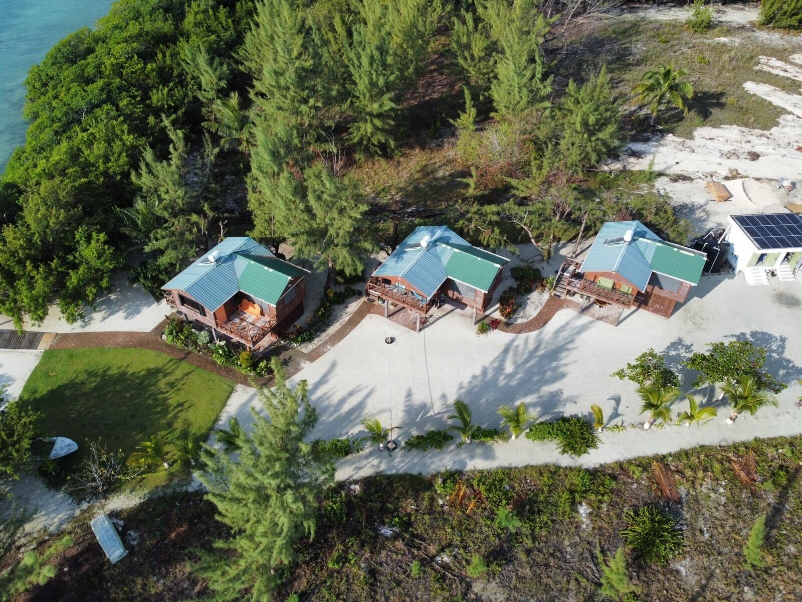 An aerial view of a coastal property with three cabins, each with a green roof, surrounded by dense greenery and a sandy beach.