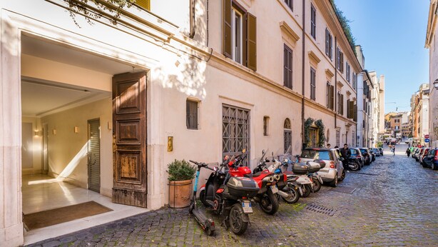 Rua de paralelepípedos com scooters e carros estacionados, entrada de prédio com porta de madeira e céu azul claro.