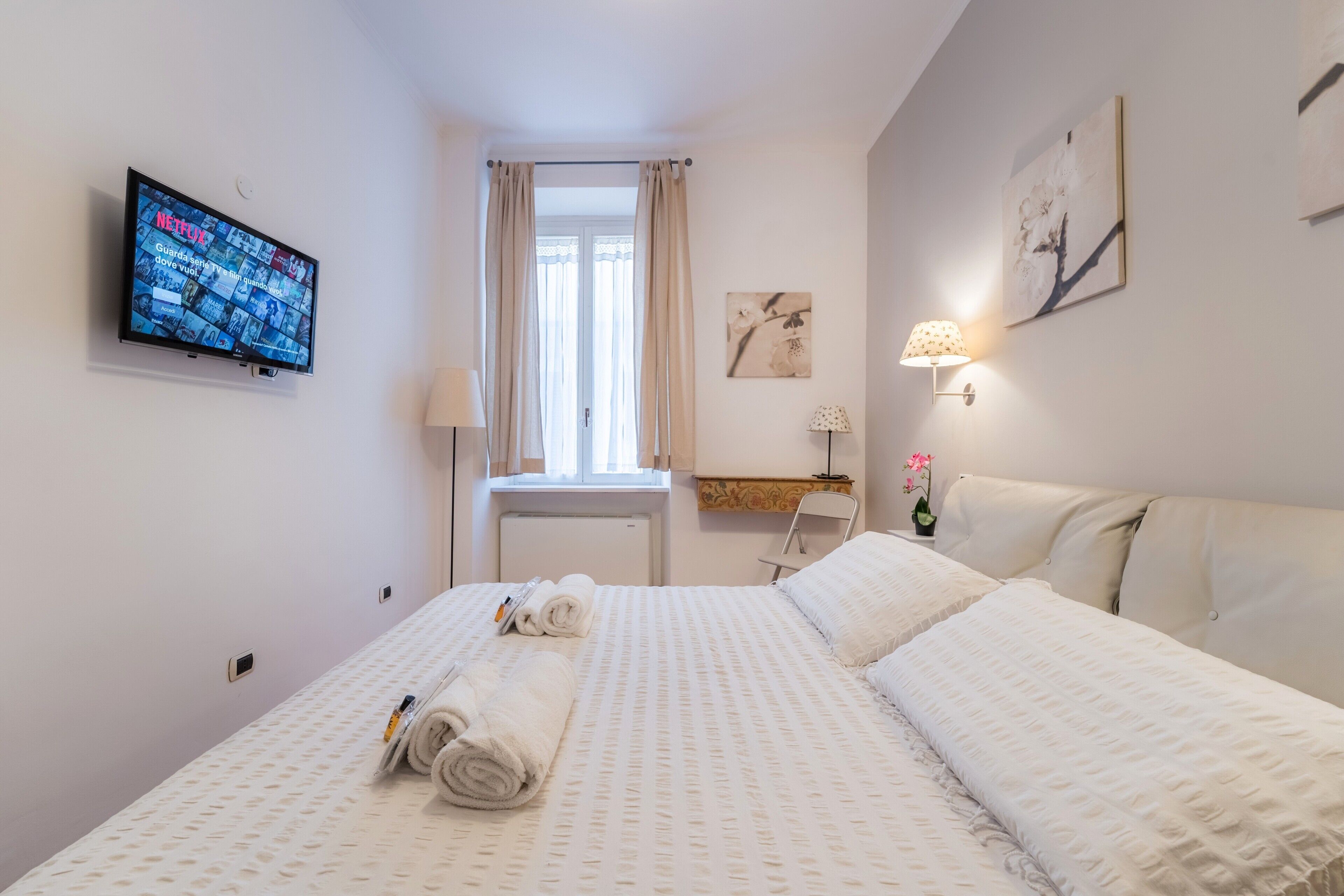 A hotel room with a neatly made bed, a television mounted on the wall, a window with curtains, and a wall-mounted lamp.