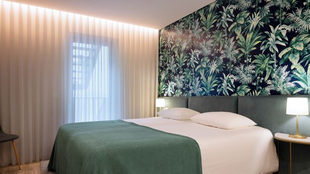Une chambre d’hôtel avec un grand lit, une table de chevet avec une lampe, une chaise et une fenêtre avec des rideaux légers.