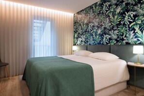 A hotel room with a large bed, a nightstand with a lamp, a chair, and a window with sheer curtains.