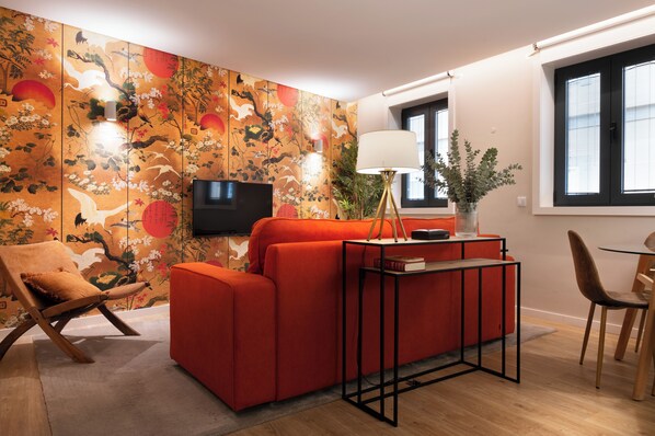 A modern living room with a red sofa, a wooden chair, a small table with a lamp, and a TV against a wall with a traditional mural.