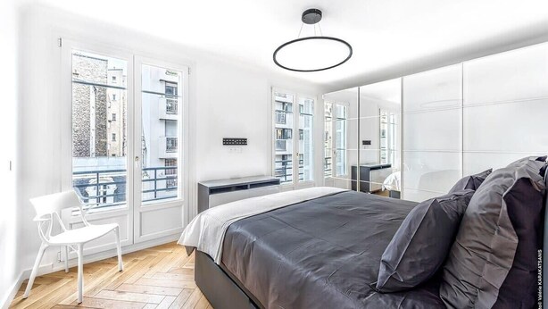 Une chambre moderne avec un grand lit, une chaise blanche et une vue sur la ville grâce à de grandes fenêtres.