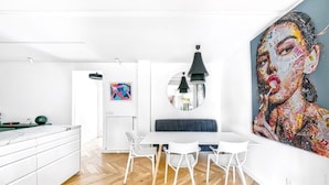Une cuisine moderne avec des meubles blancs et un parquet en bois disposé en motif chevrons. On y trouve un coin repas avec une table et des chaises blanches, ainsi qu’un grand tableau accroché au mur.