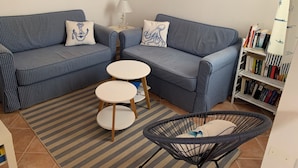 Uma sala de estar com um sofá azul xadrez, uma mesinha redonda, uma cadeira de vime, um tapete listrado e uma estante com livros.