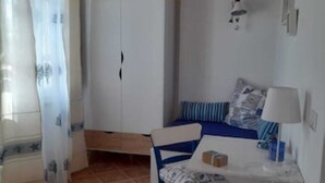 Um quarto pequeno e acolhedor com teto de madeira, um guarda-roupa branco, uma cama com roupa de cama azul, uma cadeira branca e uma pequena mesa com abajur.