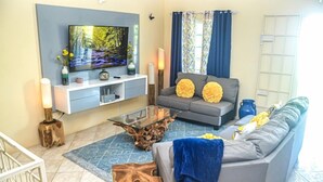 A living room with a flat-screen TV, grey sofa, decorative pillows, a glass coffee table, and a blue rug. - The Shallhome  (Montego Bay)
