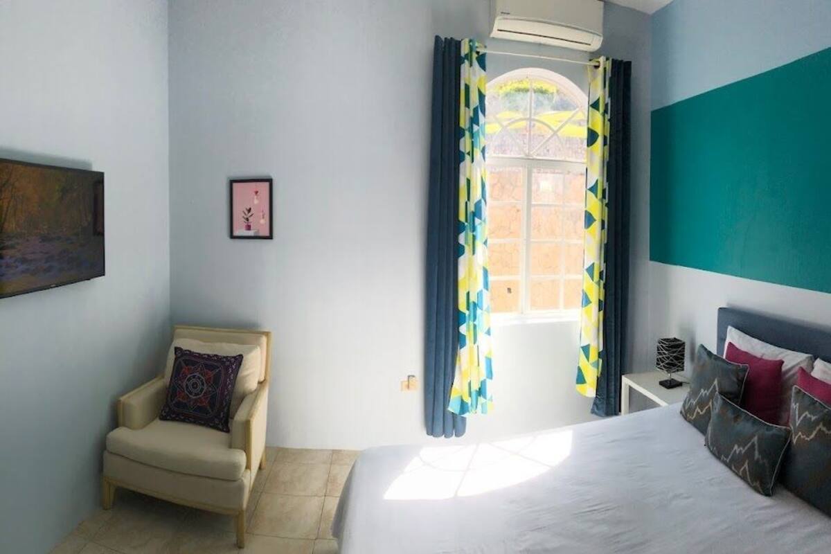 A bedroom with a bed, a chair, a window with curtains, and a painting on the wall.