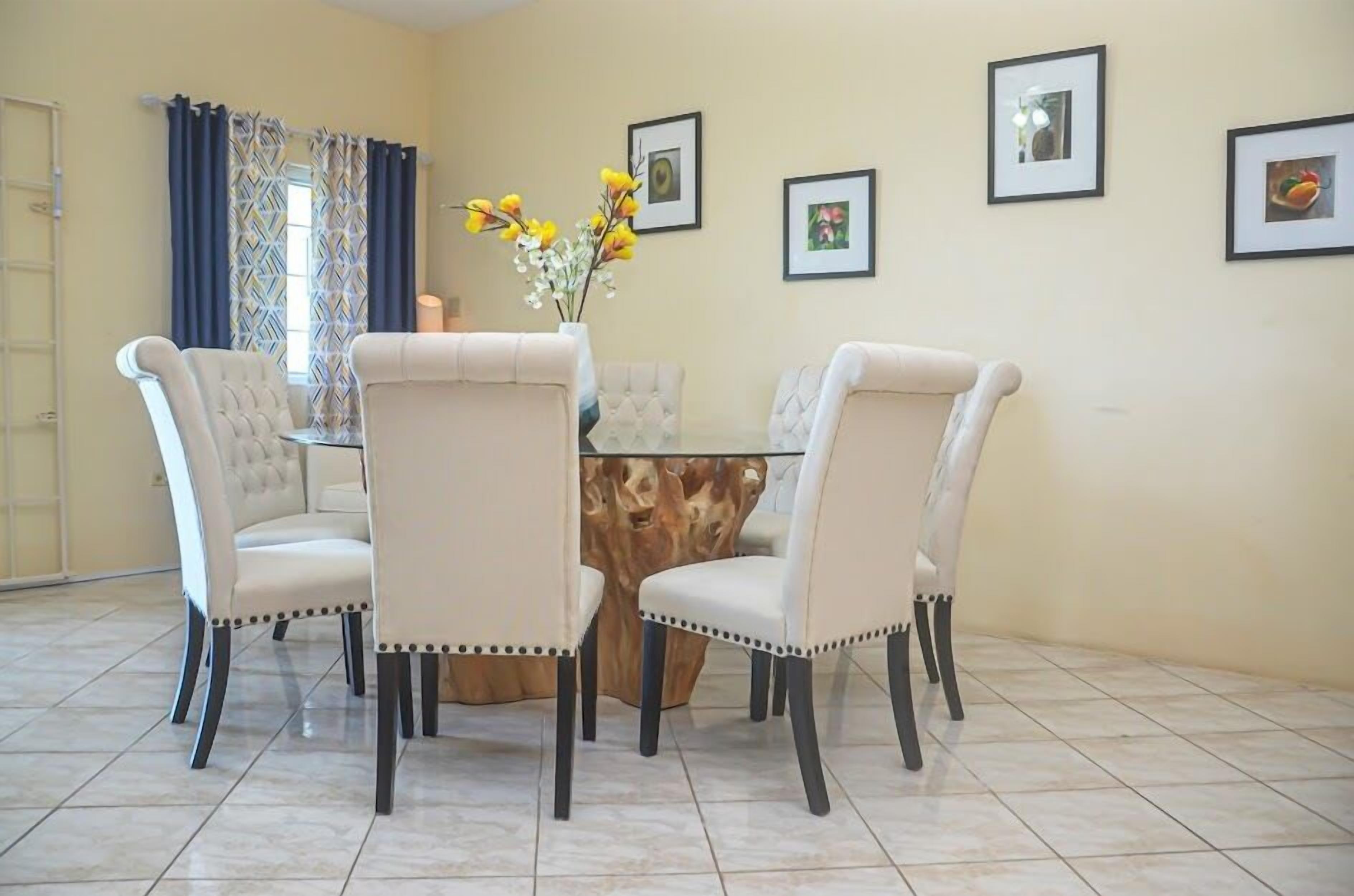 A dining area with four upholstered chairs around a round table, a vase with yellow flowers, and framed pictures on the wall.