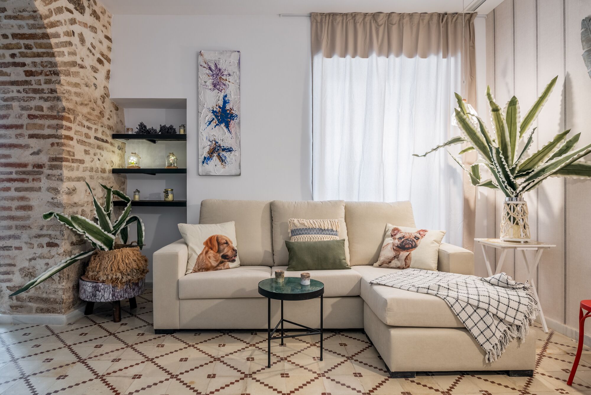 Sala de estar con sofá seccional, una mesa de centro y cojines decorativos. Hay plantas en macetas y una estantería con objetos.