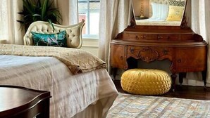 Une chambre à coucher comprenant un lit, une coiffeuse avec un miroir, une commode, une chaise et une fenêtre avec des rideaux.