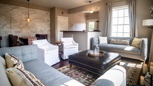 Un salon comprenant un canapé, des fauteuils, une table basse et un tapis. Un mur est décoré d’une fresque représentant un navire, une fenêtre est ornée de rideaux et un luminaire est suspendu au plafond.