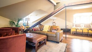 A loft-style living room with a high ceiling, exposed wooden beams, a brown leather sofa, a coffee table, and a large window providing natural light. - "Meteor": River View Penthouse in Downtown Mystic (Mystic)