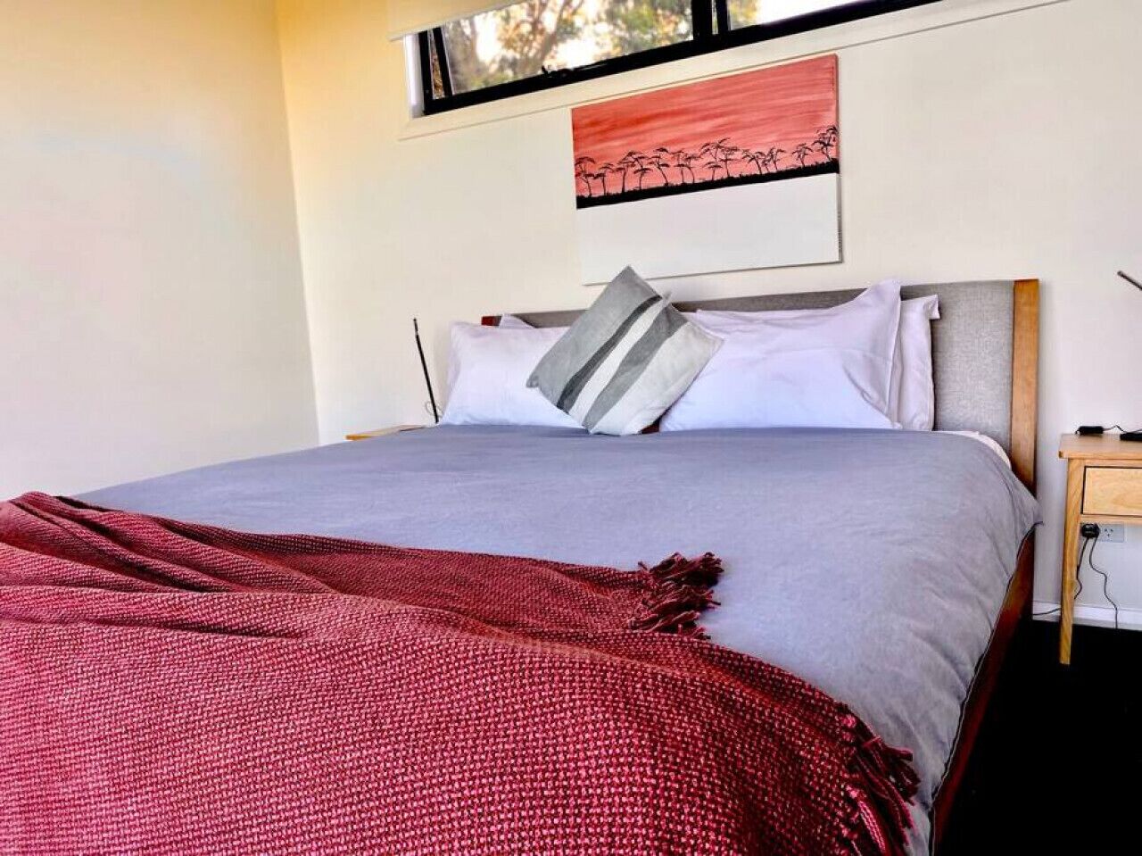 A bed with a grey and white bedding, a red blanket, and a wooden nightstand with a lamp.