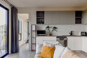 A modern kitchen with white cabinets, a built-in oven, and a dishwasher. There is a small plant on the counter and a window with curtains. - Wamoon Retreat - The Apartment ~ Luxury Gateway to Wilsons Prom (Yanakie)