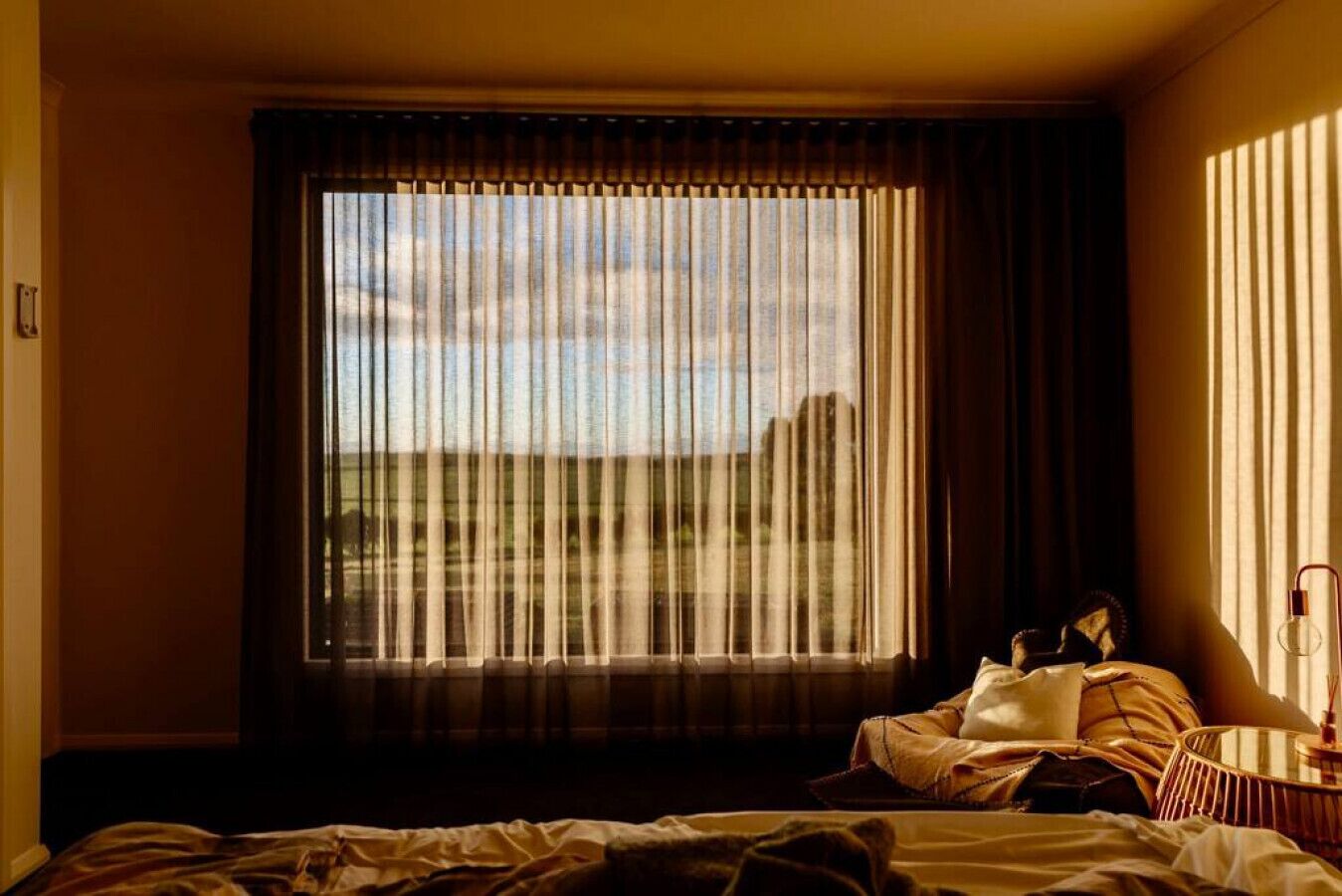 A hotel room with a large window, a bed, a bedside table, and a lamp.
