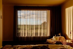 Une chambre d’hôtel avec une grande fenêtre, un lit, une table de chevet et une lampe.