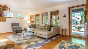 A living room with a sofa, armchair, and coffee table, wooden flooring, and large windows with blinds.