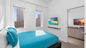 A modern bedroom with a bed, a TV mounted on the wall, and a view of the city through the windows.