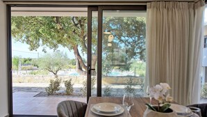 A dining area with a table set for two, a view of a pool and trees outside, and a sliding glass door. - Iris - Vacation house with private pool in Kounoupidiana, Crete (Kampáni)