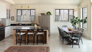 A modern kitchen with a central island, dining area with chairs, and large windows with curtains. - The Lark Haus: Luxury Home with stunning lake view (Elizabeth)