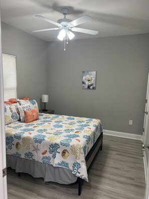 Quarto com cama, ventilador de teto, mesa de cabeceira, abajur e um quadro na parede.