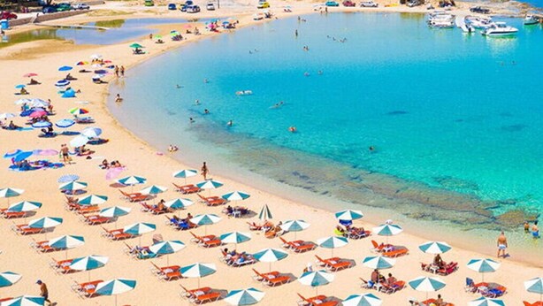 Une plage parsemée de nombreux parasols et de chaises longues, une mer d’un bleu limpide et des bateaux sur l’eau.