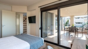 Une chambre d’hôtel moderne équipée d’un lit, d’un téléviseur fixé au mur et d’une porte-fenêtre coulissante donnant sur un balcon avec un espace salon extérieur.